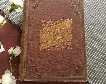 Op de afbeelding: Een vintage boek met een bruine kaft met de titel "Deportment and Social Culture" in gouden letters. De kaft heeft decoratieve hoekontwerpen. Het boek ligt op een gestreepte stof met witte bloemen.