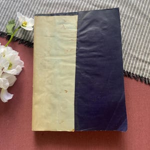 May include: A rectangular book with a two-tone cover featuring a cream-colored left side and a dark navy blue right side. The cover material appears smooth with subtle texture and slight signs of wear, including minor surface imperfections.