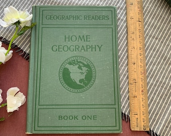 Könnte beinhalten: Ein grünes Vintage-Buch "Home Geography" aus der Reihe "Geographic Readers". Das Cover zeigt eine Weltkarte und den Text "Book One". Ein Holzlineal liegt neben dem Buch. Weiße Blumen sind im Hintergrund.