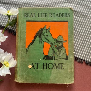 Pode incluir: Um livro de capa dura verde vintage intitulado Real Life Readers At Home. A capa apresenta uma ilustração retangular laranja central de uma pessoa com um chapéu de cowboy ao lado de um cavalo. O texto está impresso em letras pretas.