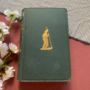 May include: A vintage, hardcover book with a dark green cover. A gold-colored illustration of a person with a staff is on the front. The book is slightly worn, suggesting age and use. The book is on a red surface.