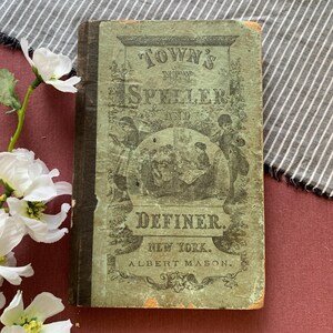 May include: An antique book titled Town's New Speller and Definer by Albert Mason, New York. The cover is light green with a dark brown spine and features a black ink illustration of children and a teacher, surrounded by decorative borders.
