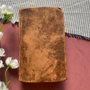 May include: A vintage, rectangular book with a worn, brown leather cover. The cover shows signs of age with visible scratches and a distressed texture. The book is positioned on a pink surface with a striped fabric and white flowers in the background.