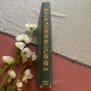 Puede incluir: Un libro verde vintage titulado "God's Calendar" con detalles florales dorados y letras. El libro est&aacute; en posici&oacute;n vertical, con flores blancas y una tela a rayas en el fondo. El nombre del autor, Quayle, y la editorial, Jennings and Graham, son visibles.