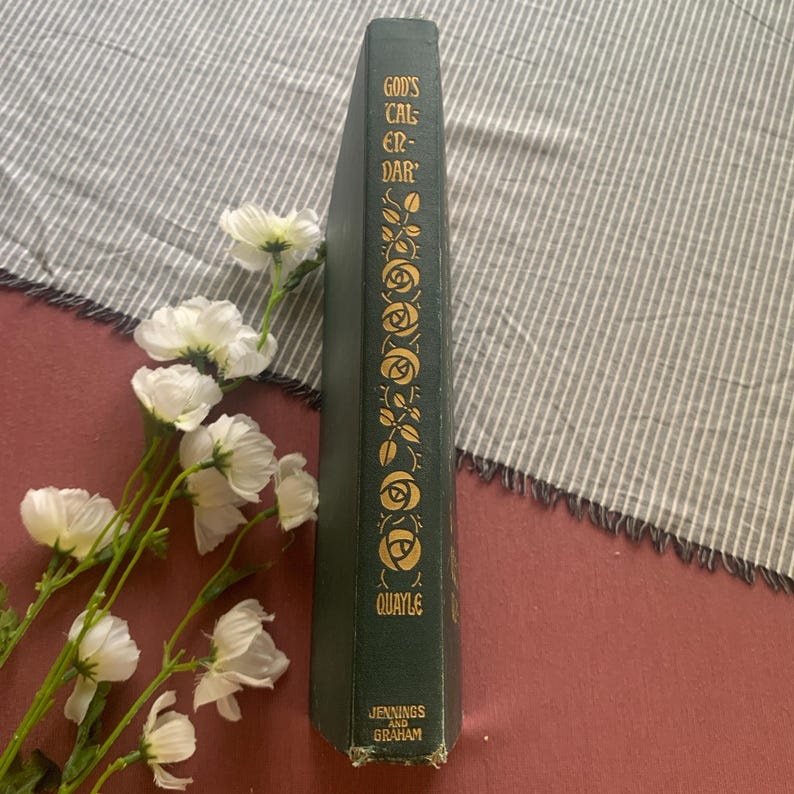 Puede incluir: Un libro verde vintage titulado "God's Calendar" con detalles florales dorados y letras. El libro est&aacute; en posici&oacute;n vertical, con flores blancas y una tela a rayas en el fondo. El nombre del autor, Quayle, y la editorial, Jennings and Graham, son visibles.