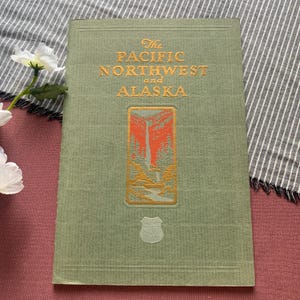 Può includere: Un libro con copertina rigida color verde salvia intitolato The Pacific Northwest and Alaska in lettere dorate. La copertina presenta un'illustrazione rettangolare di una cascata e una foresta in arancione e verde, con un piccolo stemma in rilievo.