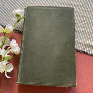 May include: An antique book with a dark green cover. The cover has a textured appearance and shows signs of wear. White flowers with green stems are in the corner of the image. The book is on a red surface with a striped cloth.