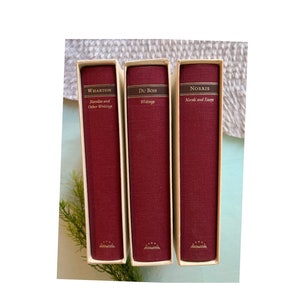 Puede incluir: Tres libros de tapa dura rojos con letras doradas en el lomo. Los títulos son "Wharton Novels and Other Writings", "Du Bois Writings" y "Norris Novels and Essays".