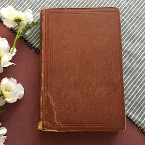 May include: A vintage hardcover book with a textured, brown cover. The book's edges show signs of wear, suggesting age. White flowers with green stems are arranged on a burgundy surface, with a striped gray and white fabric in the background.