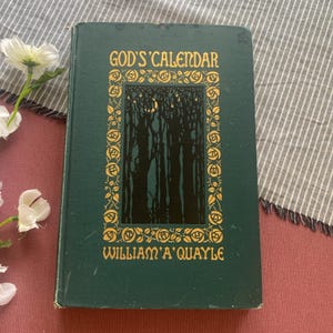 Puede incluir: Un libro antiguo de tapa dura titulado "God's Calendar" de William A. Quayle. La portada es de color verde oscuro con letras doradas y una ilustraci&oacute;n de una escena de bosque. El libro est&aacute; sobre una superficie roja con flores blancas.