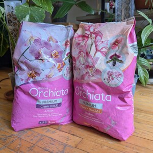 May include: Two pink bags of "Orchiata" orchid substrate. The bags feature images of orchids and text indicating "Premium" quality and different particle sizes, with one bag labeled "Classic" and the other "Power+".