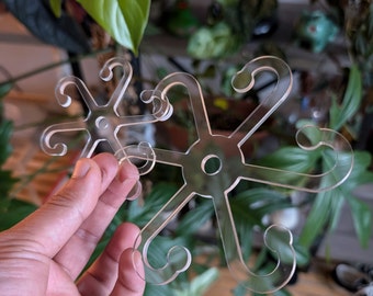May include: Two clear plastic snowflake-shaped ornaments with a circular center and six curved arms. The ornaments are held in a hand.