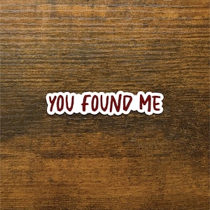 May include: A white sticker with the words "YOU FOUND ME" in a bold, maroon font. The sticker is rectangular and placed on a brown wooden surface. The text is centered and easy to read.