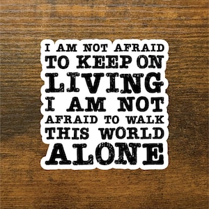 May include: White sticker with black text that reads: "I AM NOT AFRAID TO KEEP ON LIVING I AM NOT AFRAID TO WALK THIS WORLD ALONE." The sticker has a black border and is set against a wooden background.