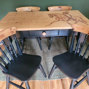 May include: A wooden dining table with a black base and a light wood top featuring a horse and rider design. Two black chairs with matching horse designs on the backrests are in front. Two additional chairs are behind the table.
