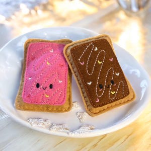 Puede incluir: Dos pasteles de fieltro, que se asemejan a pasteles tostados, uno rosa y otro marrón, descansan sobre un plato blanco. El pastel rosa tiene glaseado rosa con detalles de corazón y amarillo. Ambos tienen caras sonrientes.