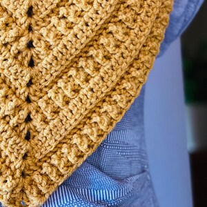 May include: A close-up of a crocheted scarf in a mustard yellow colour. The scarf is made with a simple stitch pattern and has a triangular shape.