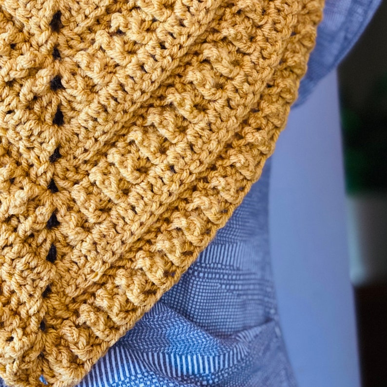 May include: A close-up of a crocheted scarf in a mustard yellow colour. The scarf is made with a simple stitch pattern and has a triangular shape.
