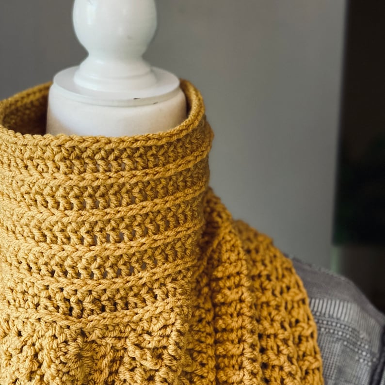 May include: A crocheted neck warmer in a mustard yellow colour. The neck warmer is made with a simple stitch pattern and is shown on a white mannequin.