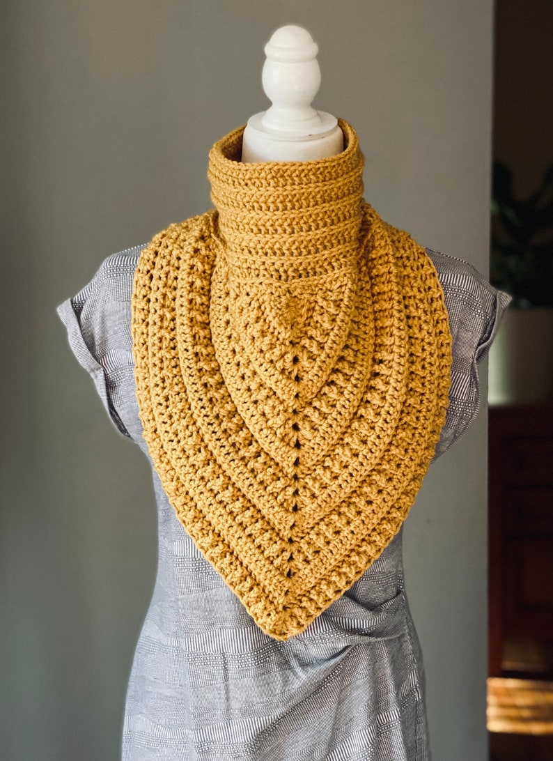 May include: A crocheted cowl in a mustard yellow colour. The cowl is made with a simple stitch pattern and has a triangular shape.