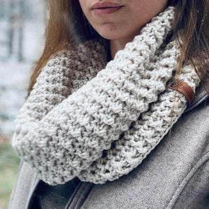 Könnte beinhalten: Ein grob gestrickter Infinity-Schal in hellem Cremeweiß mit einem strukturierten, erhabenen Strickmuster. Der Schal ist mit einem kleinen, rechteckigen braunen Lederetikett an der Seite versehen, auf dem der Text "CHARLIER" eingeprägt ist.