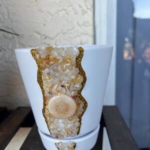 May include: White ceramic plant pot with a decorative design. The design features a vertical strip of small, light-colored stones, outlined with gold glitter. A round, light-colored stone with a symbol is centered on the pot.