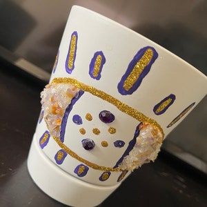 May include: White ceramic planter with a purple and gold eye design. The eye is decorated with small, clear crystals and purple gems.