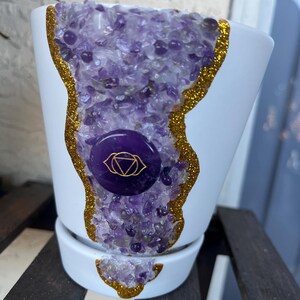 May include: White ceramic planter with a gold glitter rim and a purple amethyst crystal design. The planter features a purple and gold symbol of the third eye chakra.