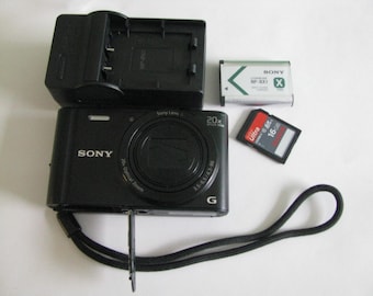 May include: A black Sony digital camera with a 20x optical zoom lens, a black battery charger, a silver Sony NP-BX1 lithium-ion battery, and a black SanDisk Ultra 16GB SD memory card. The camera includes an attached black wrist strap.
