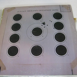 Puede incluir: Una hoja de práctica de tiro vintage con múltiples dianas negras sobre un fondo lila claro. La hoja está etiquetada como "JULIAN BREAM'S GREATEST HITS" y muestra signos de uso con agujeros de bala.