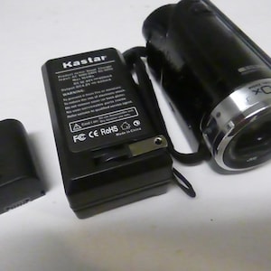 Pode incluir: Uma câmara de vídeo preta com uma lente prateada e um zoom de 40x. A câmara de vídeo está ao lado de um carregador de bateria preto e uma bateria preta.