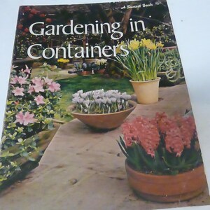 May include: A book cover with the title "Gardening in Containers" in a white font on a green background. The cover features a variety of plants in pots, including pink flowers, white flowers, and yellow flowers.