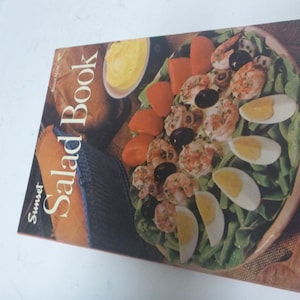 May include: A vintage cookbook titled "Sunset Salad Book" featuring a colourful image of a salad with shrimp, olives, tomatoes, and hard-boiled eggs.