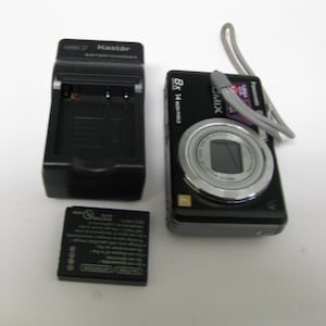 May include: A black digital camera with a silver lens, a black battery charger, and a black battery are displayed. The camera has a wrist strap. The charger is labeled "Kastar Battery Charger."