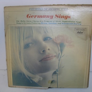 May include: Vintage vinyl record album cover titled "Germany Sings." The cover features a woman with blonde hair and blue eyes, holding a pink flower. The album title is in red, with additional text in blue.
