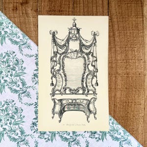 May include: An antique print of a furniture design, featuring an ornate table with a large mirror and draped fabric. The black and white illustration showcases intricate details and a classic aesthetic. The text at the bottom reads "Design for a Toilet Table."