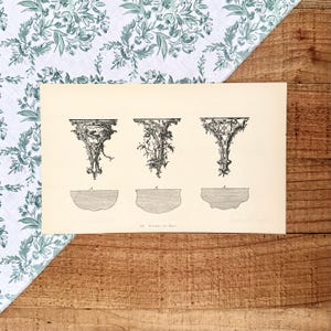 May include: An antique print featuring three ornate bracket designs. The brackets are intricately detailed with scrolling patterns and decorative elements. The print is on aged paper, with the title "66. Brackets for Busts" at the bottom. The background includes a floral patterned fabric and a wooden surface.