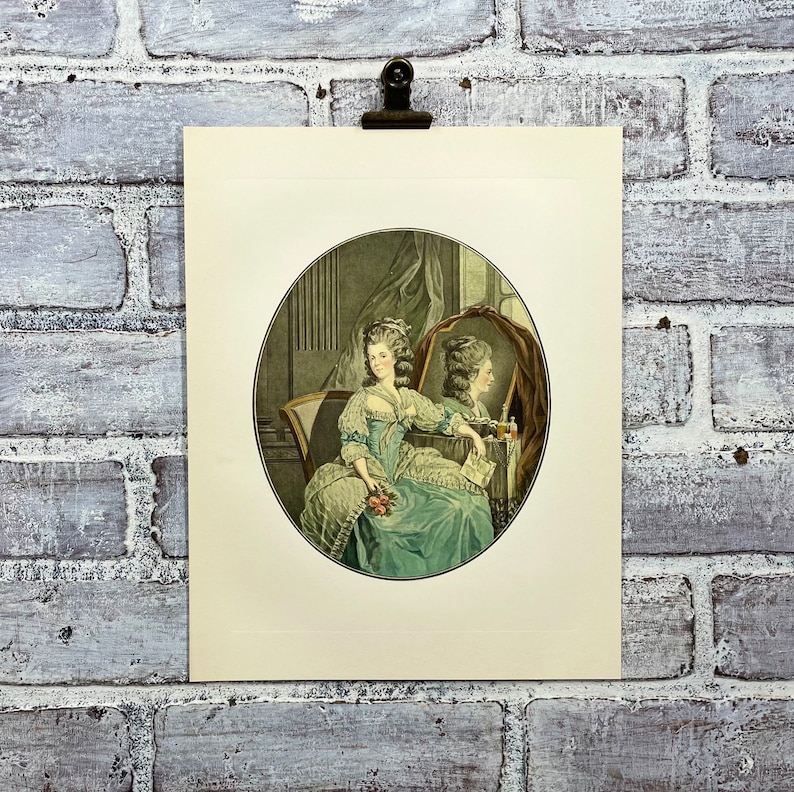 May include: A vintage print of a woman in a blue and white dress sitting in a chair and looking at her reflection in a mirror. The woman is holding a bouquet of flowers.