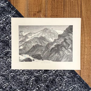 May include: Black and white mountain landscape print. The print features a detailed illustration of a mountain range with snow-capped peaks. The print is on a cream-colored paper and is set against a black and white paisley patterned fabric and a wooden surface.