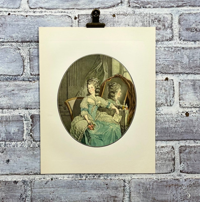 May include: A vintage print of a woman in a blue and white dress sitting in a chair and looking at her reflection in a mirror. The woman is holding a bouquet of flowers.