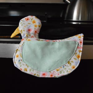 May include: A duck-shaped oven mitt with a floral pattern in yellow, pink, and blue on a white background. The mitt has a light green pocket and a yellow beak. The mitt is sitting on a black stove top.