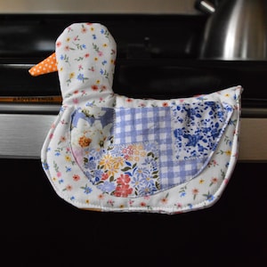 May include: A duck-shaped oven mitt with a patchwork design. The mitt features a white base with floral patterns, a blue gingham check, and a blue floral print. The duck's beak is orange with white polka dots.