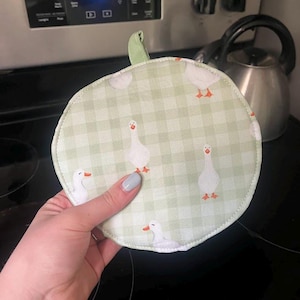 May include: Round, light green and white gingham patterned pot holder with white geese illustrations. The pot holder has a green loop for hanging. The pot holder is approximately 7 inches in diameter.