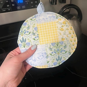 May include: Round quilted pot holder with a patchwork design. The pot holder features a mix of floral and gingham patterns in blue, yellow, and white. It has a small loop for hanging.