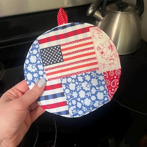 May include: Round, patriotic-themed pot holder featuring a patchwork design with the American flag, blue floral patterns, and red and white stripes. It has a small red loop for hanging.