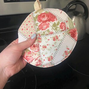 May include: A round, quilted pot holder with a floral pattern in shades of red, pink, and green on a white background. The pot holder has a loop for hanging and is made of fabric. Kitchenware.