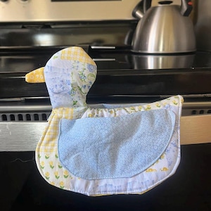 May include: A quilted duck-shaped oven mitt with a yellow, blue, and white floral pattern. The mitt has a blue center and a yellow beak. The mitt is designed to protect hands from heat when handling hot cookware.