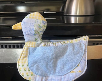Puede incluir: Un guante de cocina acolchado con forma de pato con un estampado floral amarillo, azul y blanco. El guante tiene un centro azul y un pico amarillo. Está diseñado para proteger las manos del calor al manipular utensilios de cocina calientes.