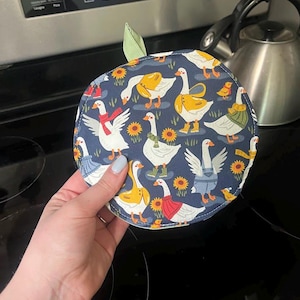 May include: A round, navy blue pot holder with a whimsical goose pattern. The geese are depicted in various outfits, surrounded by sunflowers and rain boots. A green leaf is attached to the top.