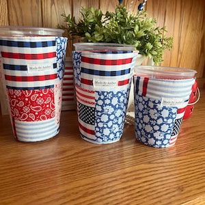 May include: Three cylindrical fabric cup sleeves in varying sizes, each featuring a patchwork design of red, white, and blue patterns including gingham, paisley, and floral prints. Each sleeve has a white label reading Made By Amber.